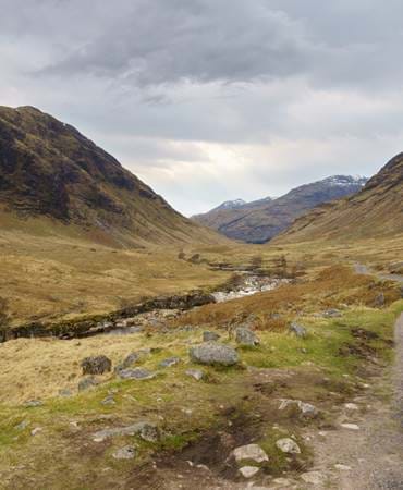 A rocky path through a valley with mountains in Perthshire looming in the distance, under dramatic skies.