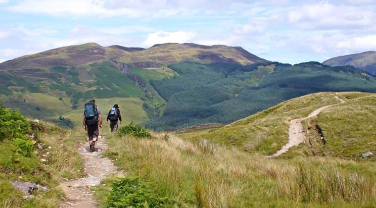 Two hikers with backpacks walk a grassy trail toward forested hills under a partly cloudy sky in Scotland.