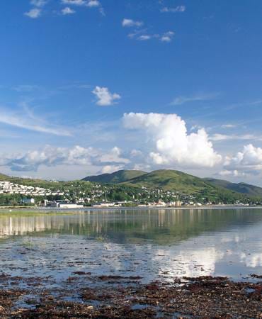 Scenic coastal Scottish seaside town with white houses nestled between green hills and reflective water.